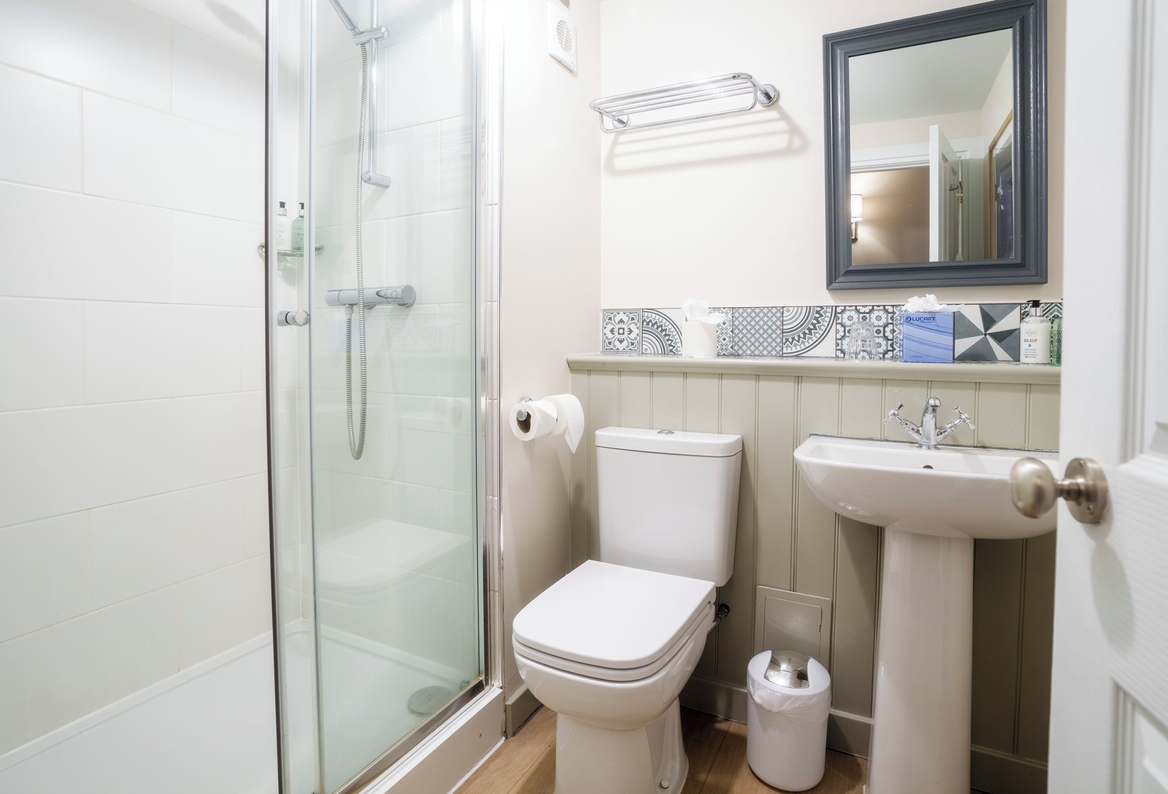 Un baño con ducha envuelta en vidrio, un inodoro blanco, un lavamanos pedestal y un espejo sobre él.