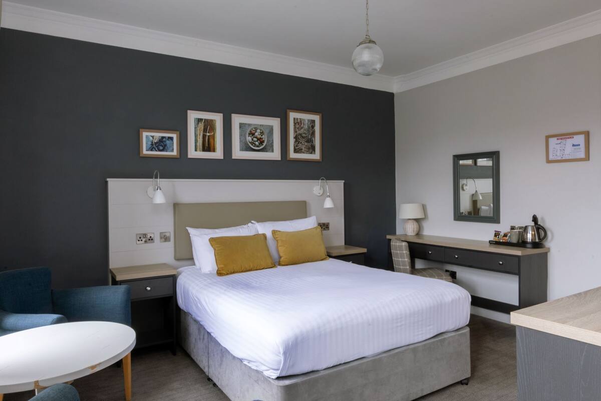 A modern hotel room with a large bed, bedside tables, a desk, and a chair. There are framed pictures on the wall and a mirror above the desk.