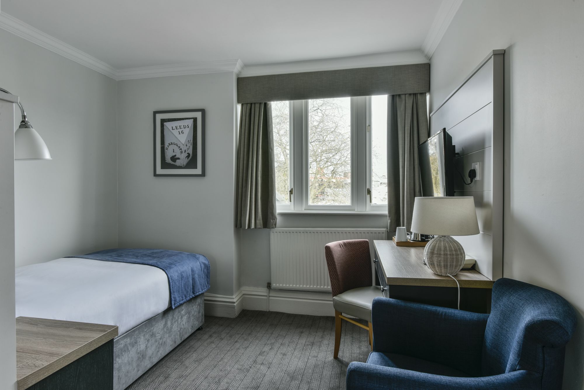 A hotel room with a bed, a desk with a lamp, a chair, a window with curtains, and a framed picture on the wall.
