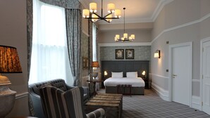 Habitación de hotel con una cama grande, un área de descanso con una silla y una mesa, un candelabro y vistas al exterior a través de una ventana con cortinas.