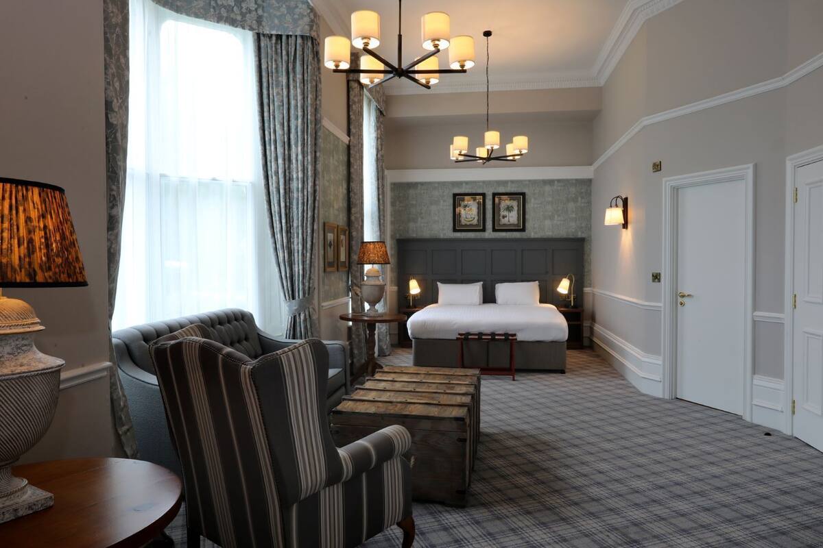 A hotel room with a large bed, a sitting area with a chair and a table, a chandelier, and a view of the outside through a window with curtains.