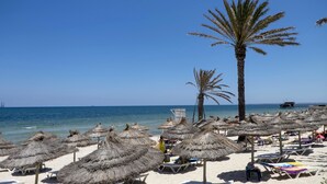 Una playa con sombrillas de techo de paja, sillas de descanso y palmeras.