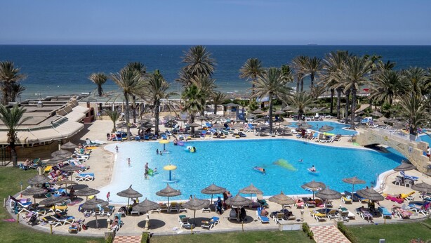 Una gran piscina al aire libre rodeada de palmeras y sillas de descanso, con vista al mar.