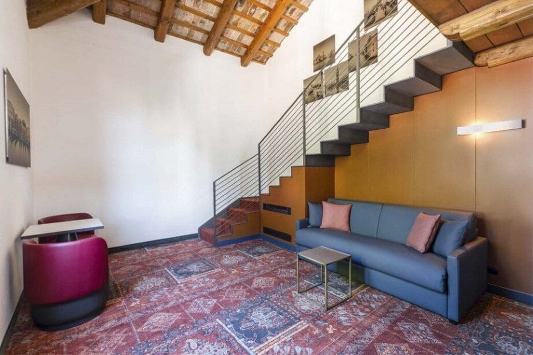 A modern living room with a blue sofa, a red and black chair, a small table, and a staircase with a wooden finish.