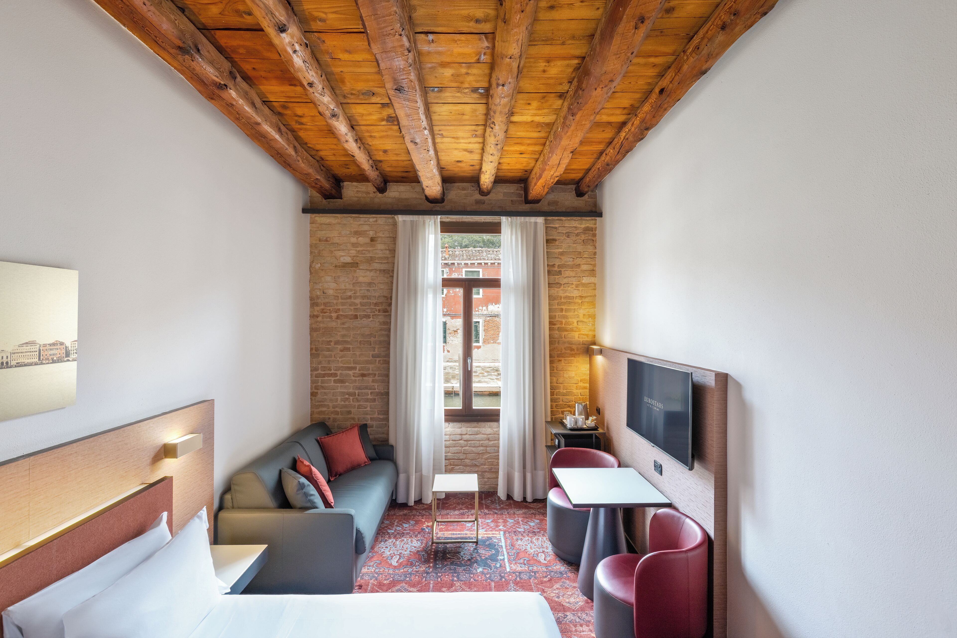 A hotel room with a wooden ceiling, a brick wall, a sofa, a bed, a TV, and a small table.