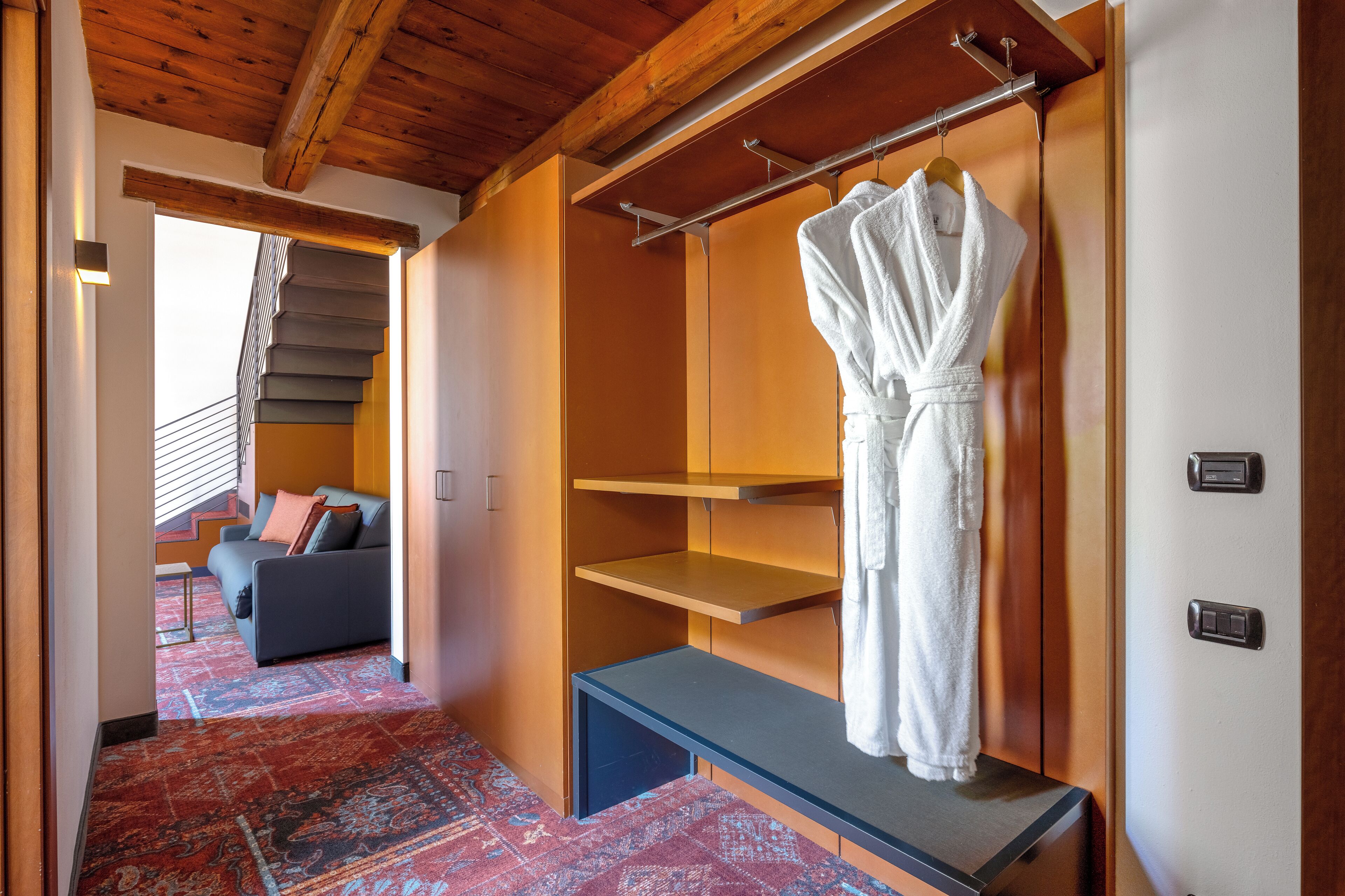 A hotel room with a wooden ceiling, a staircase, a sofa, and a hanging robe.