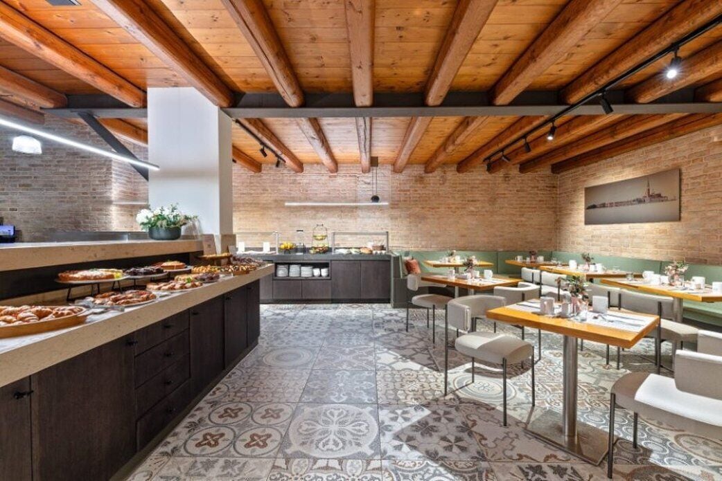 A restaurant with a rustic wooden ceiling, a brick wall, and a tiled floor. There are tables and chairs arranged for dining.