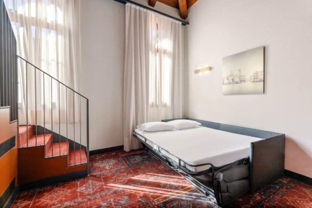 Habitación pequeña con cama, un escritorio, una silla, una escalera y una ventana con cortinas.