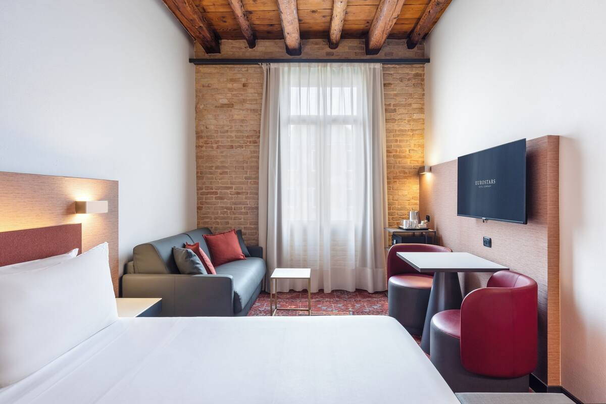 A hotel room with a bed, a sofa, a small table, a TV, and a brick wall.