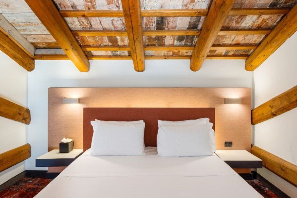 A bedroom with a wooden ceiling, a large bed with white bedding, two bedside tables, and a nightstand with a tissue box.