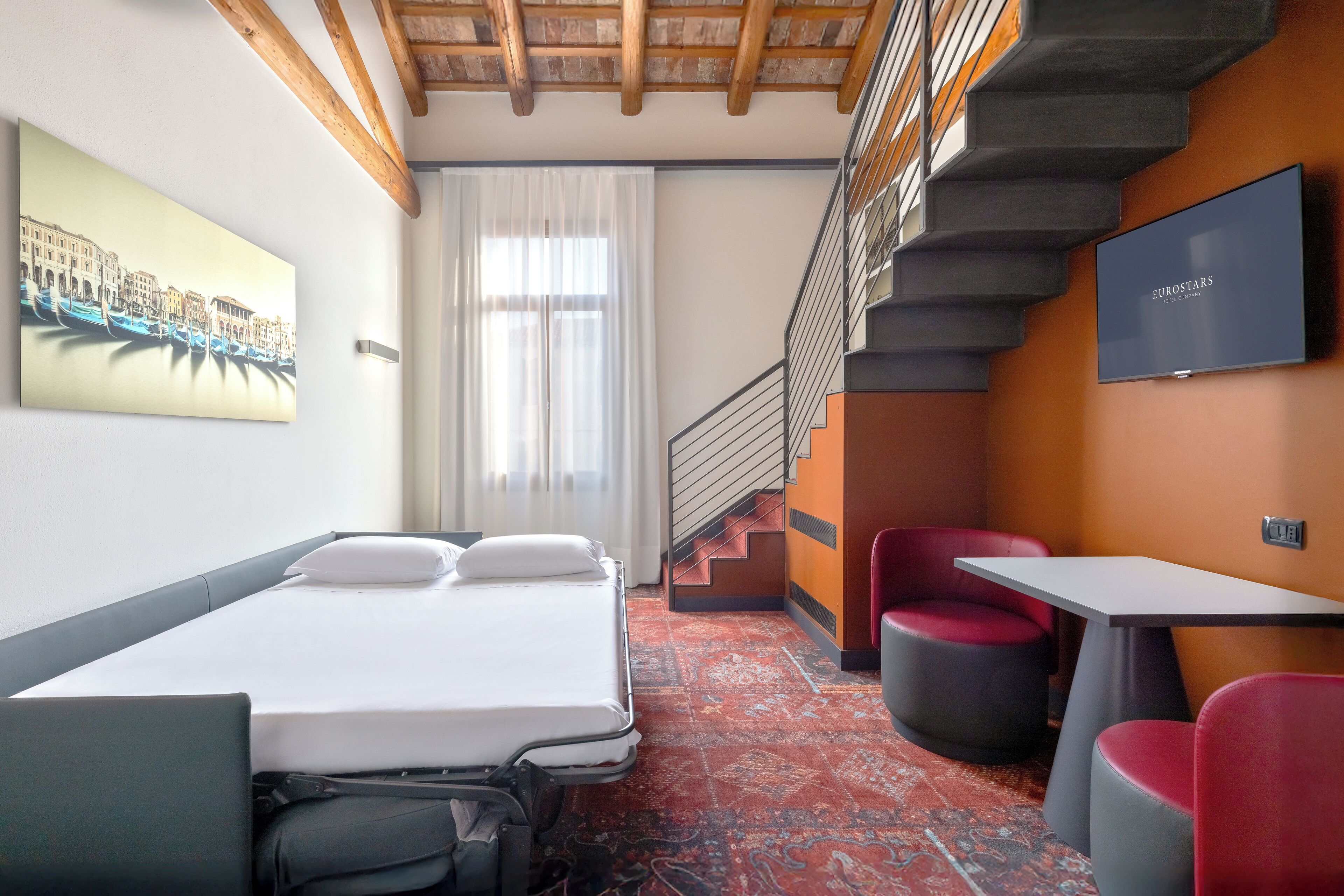 A hotel room with a bed, a desk, a chair, a TV, a staircase, and a painting on the wall.