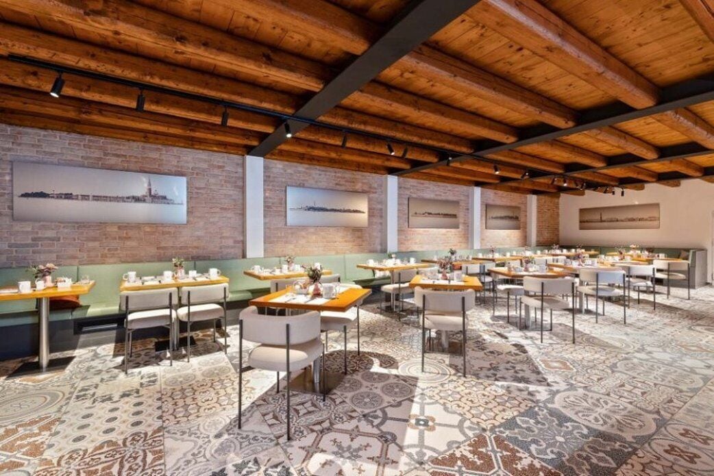 Un restaurant avec des poutres en bois au plafond, des murs en briques et une moquette à motifs. Les tables et les chaises sont disposées pour accueillir les clients.