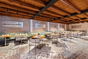 Un ristorante con travi in legno al soffitto, pareti in mattoni e un tappeto decorato. Tavoli e sedie disposti per consumare un pasto.