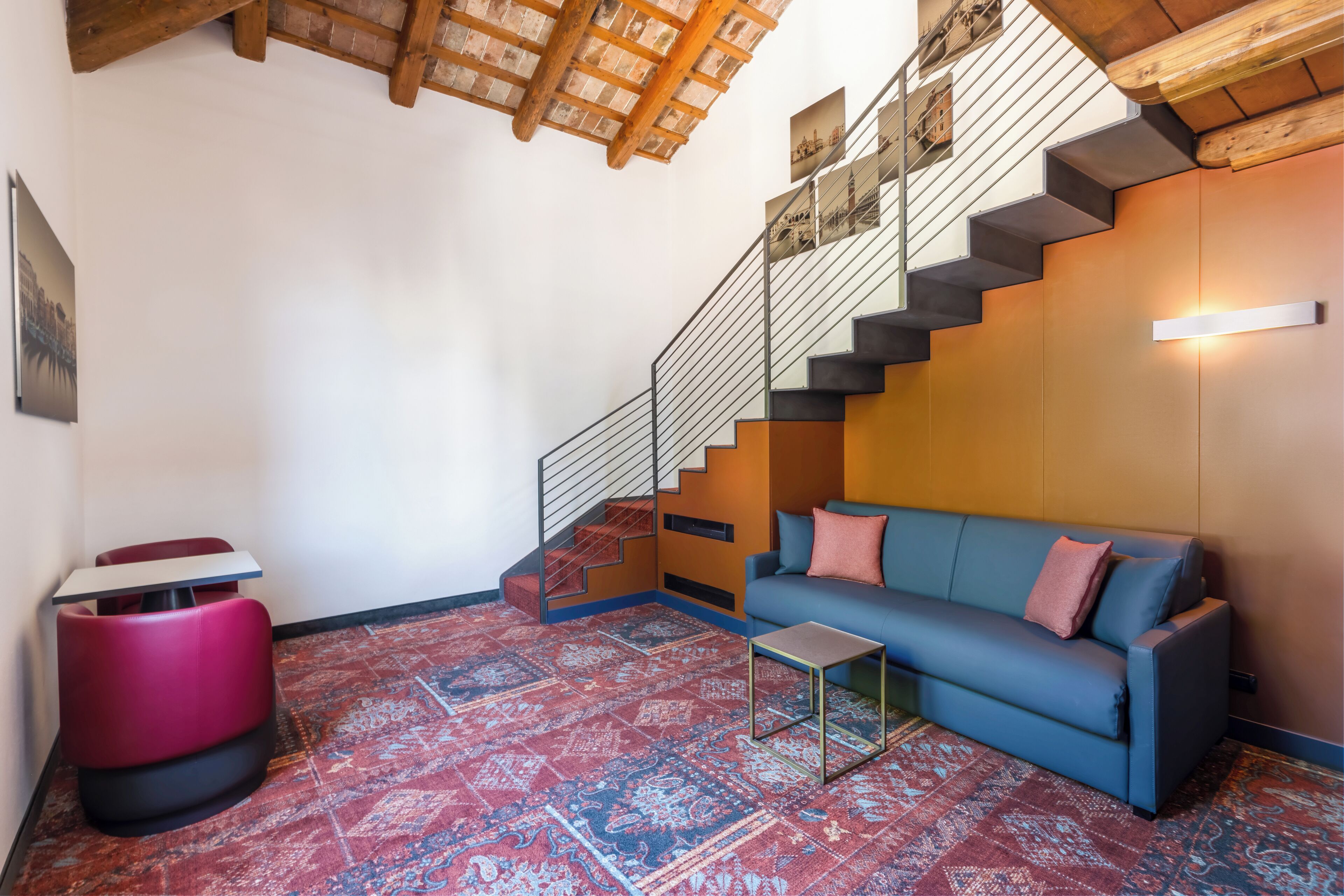 A modern living room with a blue sofa, a red ottoman, a small table, a staircase with a metal railing, and a patterned rug.