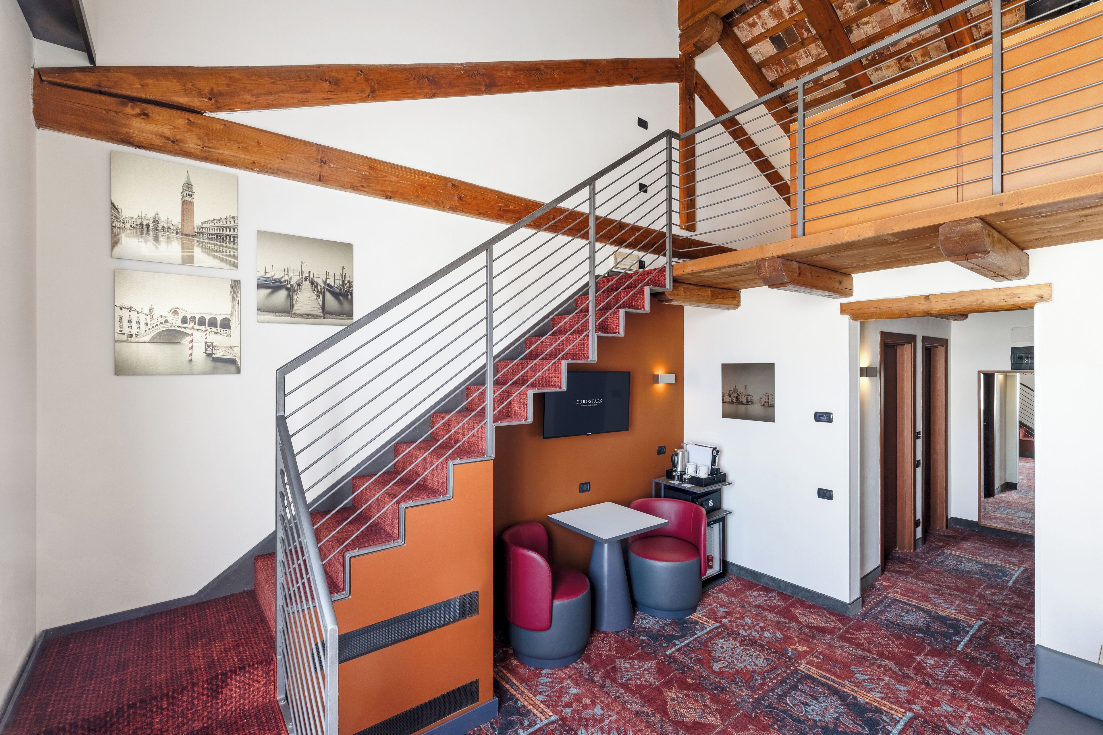 A staircase with a metal railing leading to a mezzanine, a small table with two chairs, and a patterned carpet.