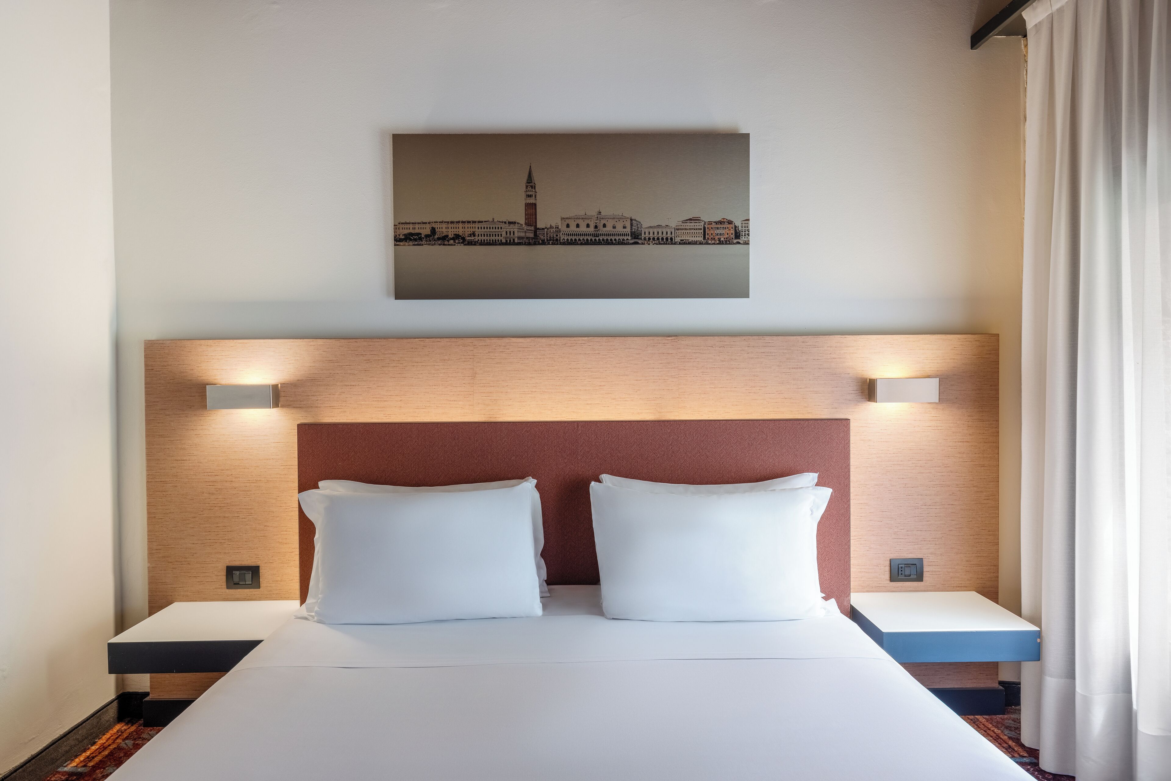 Een hotelkamer met een groot bed, twee nachtkastjes, een hoofdbord met verlichting en een stadslandschap schilderij erboven.
