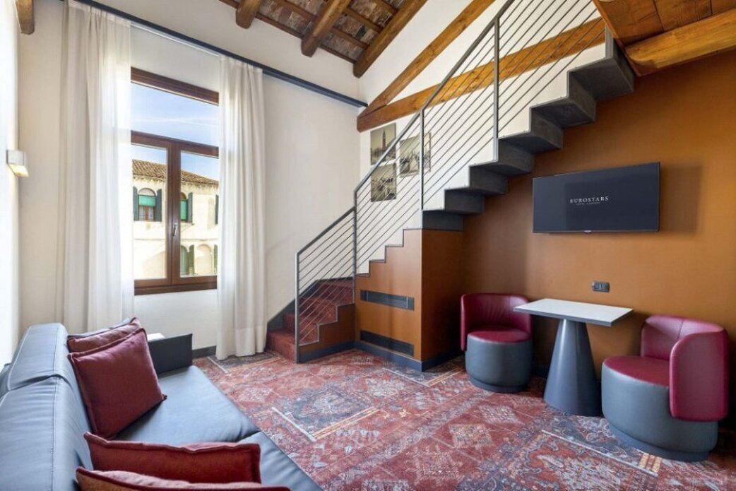 A modern hotel room with a sofa, two chairs, a small table, and a staircase.