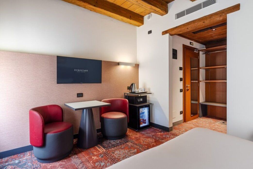Habitación de hotel con una cama, dos sillas rojas y negras, una mesita, un televisor montado en la pared y un mini bar.