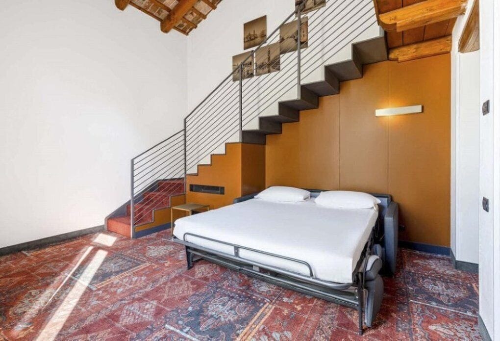 A compact room with a bed, a small bedside table, a patterned rug, a staircase with a railing, and a wall-mounted light.
