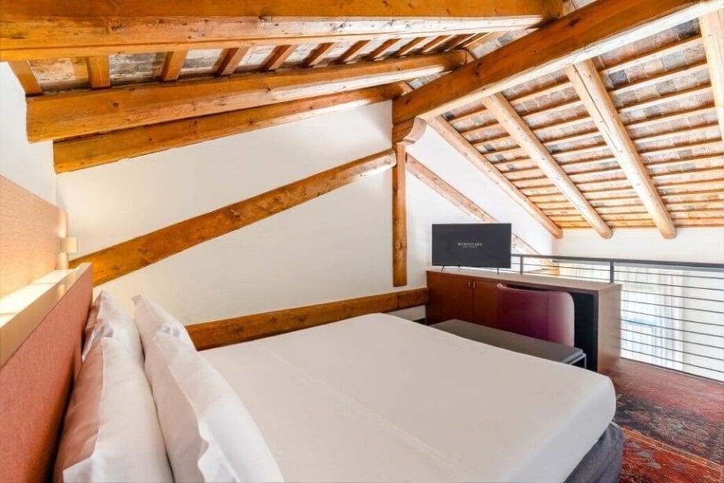 A bedroom with a sloped ceiling, wooden beams, a bed with white linens, a TV, and a view of the outdoors.