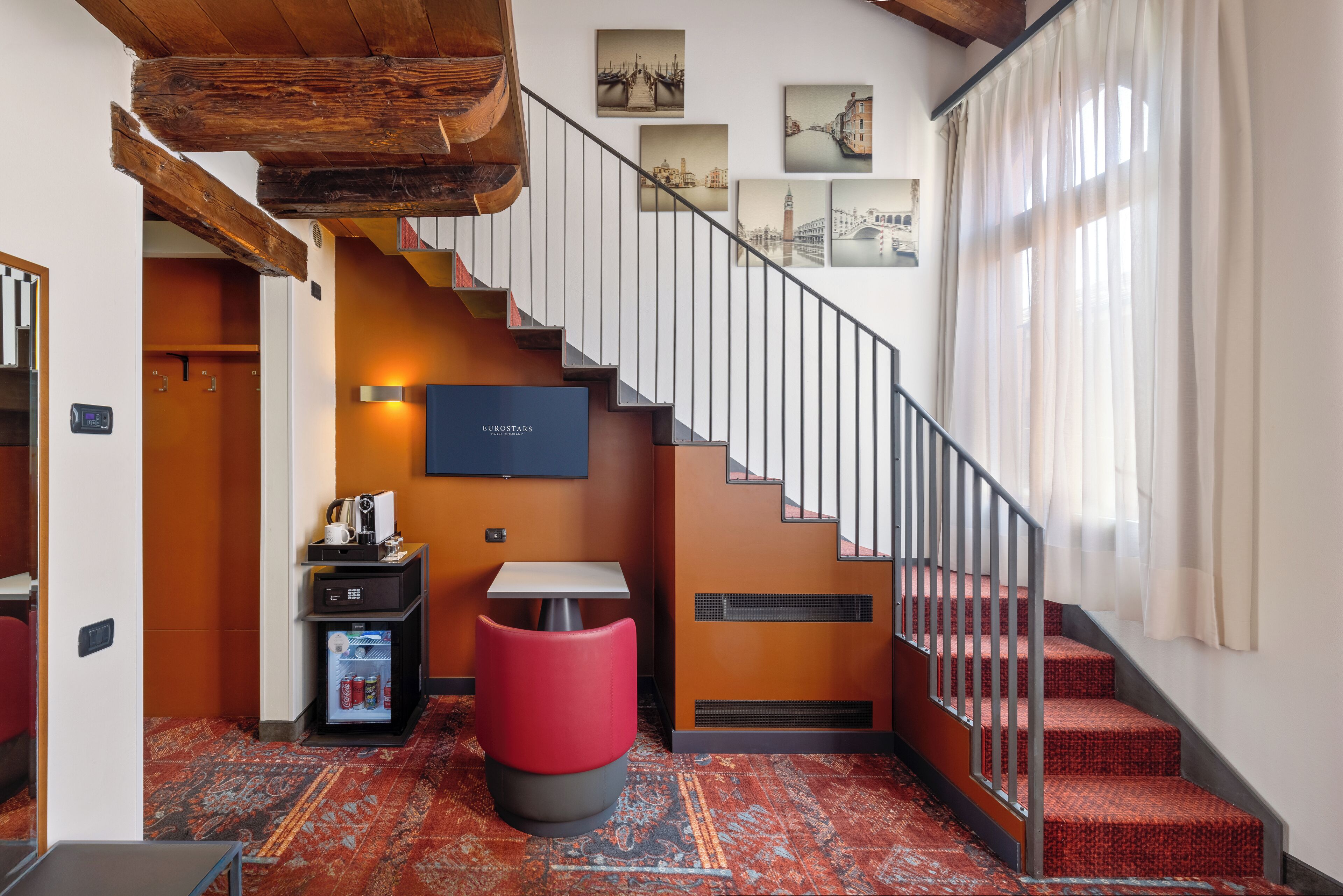 A hotel lobby with a staircase, a red ottoman, a minibar, and framed pictures on the wall.