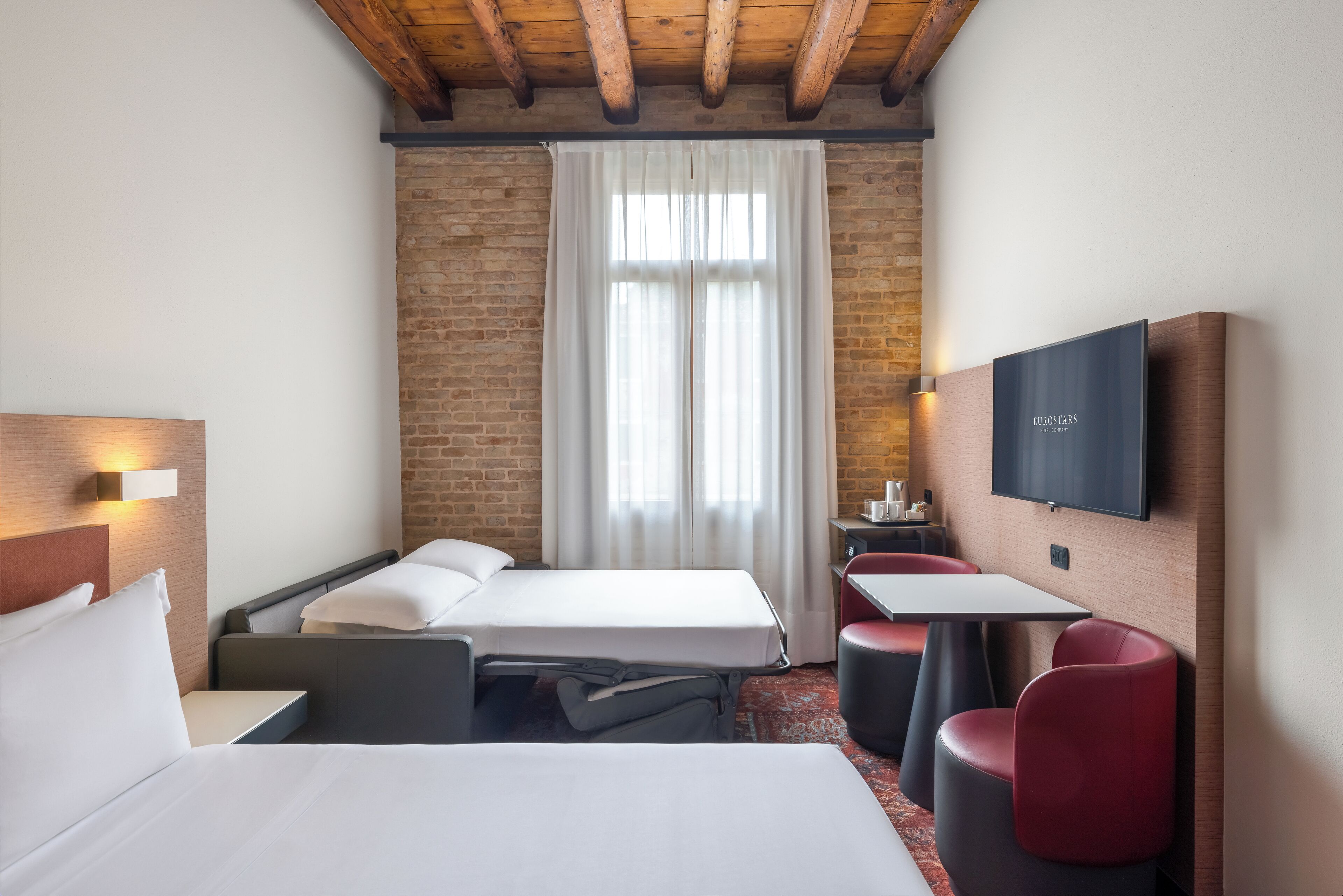 A hotel room with two beds, a TV, a small table, and a red chair.