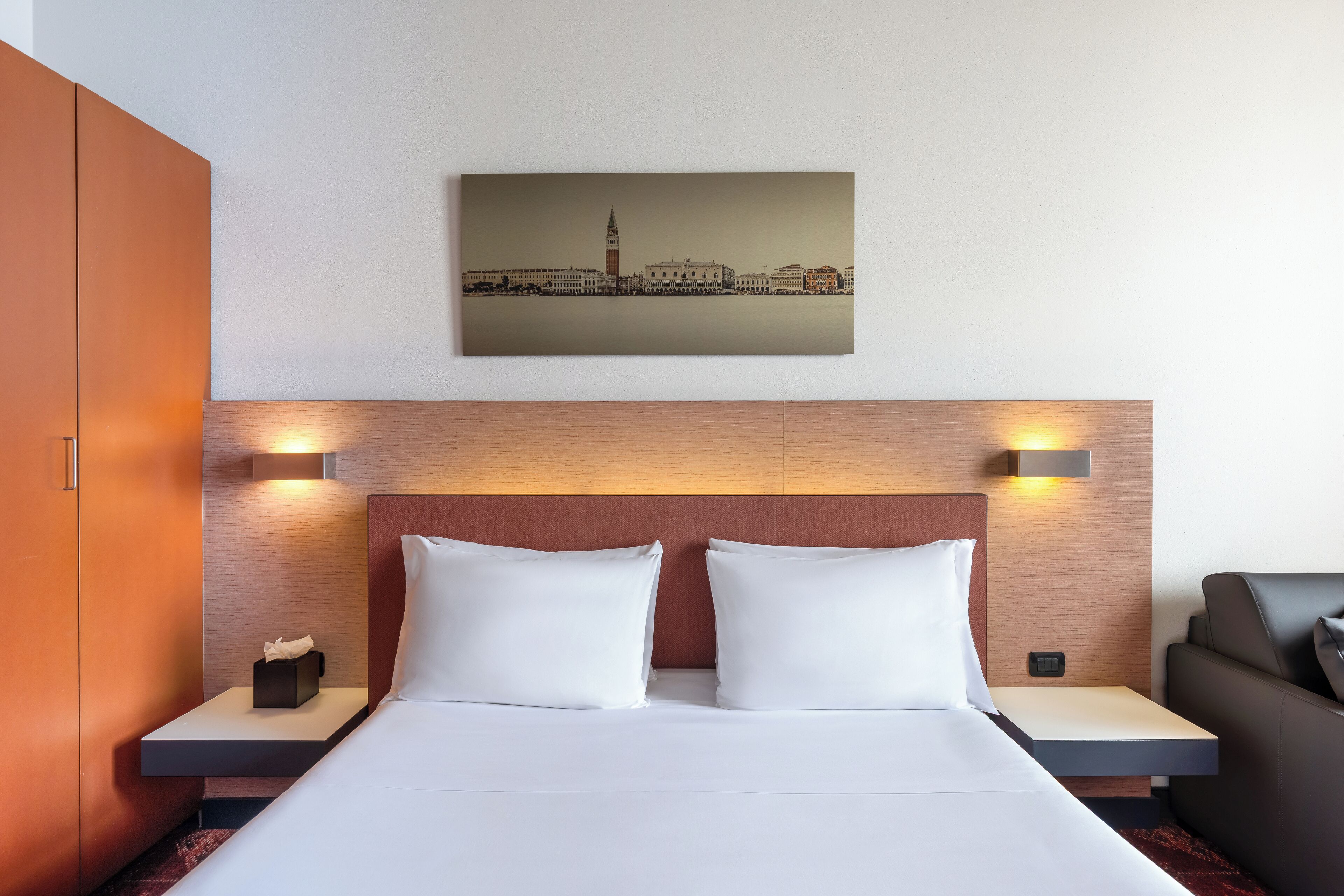 Une chambre d’hôtel comprenant un lit, des tables de chevet, une petite table, une tête de lit et une œuvre d’art fixée au mur représentant un paysage urbain.