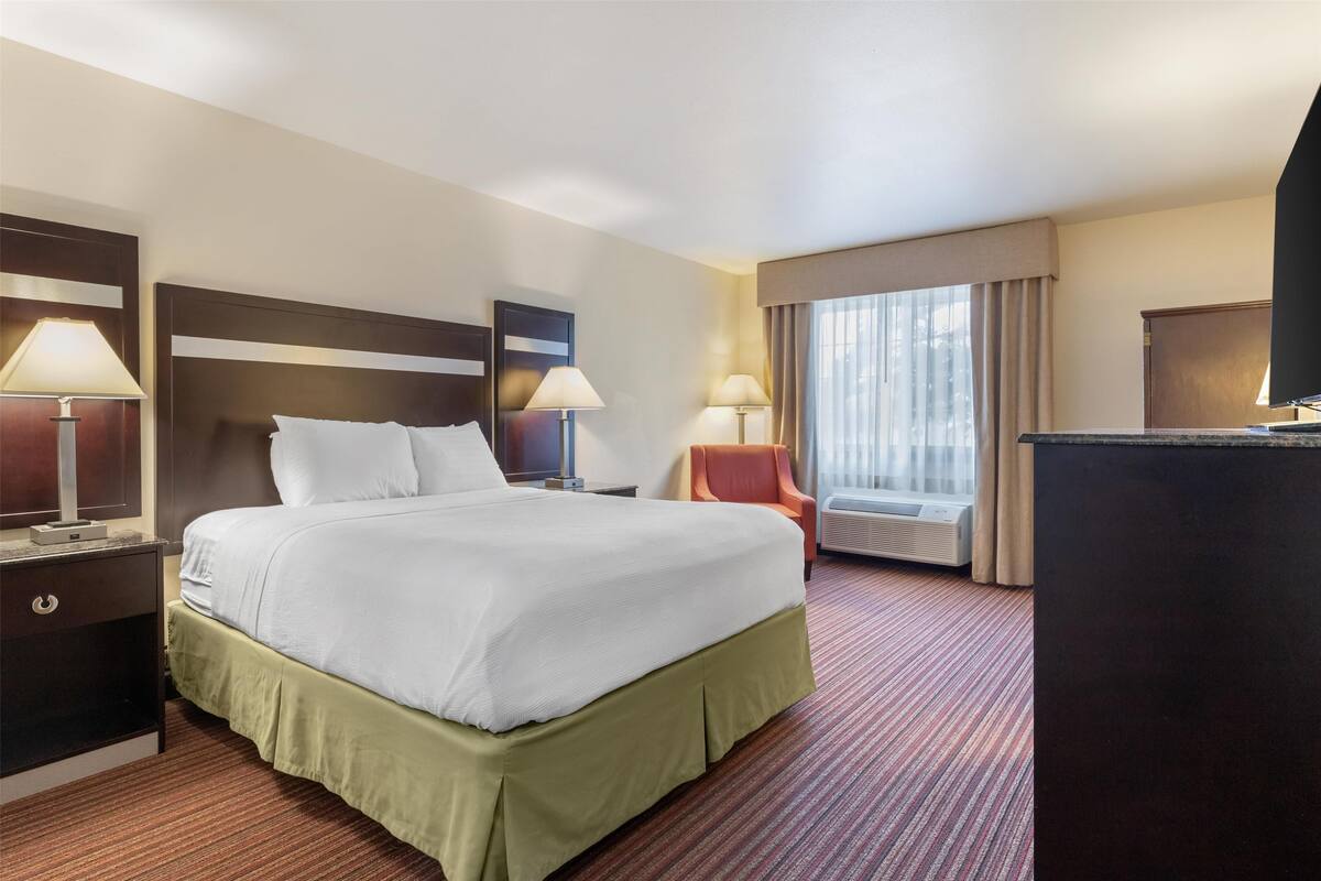 Une chambre d’hôtel avec un grand lit, deux tables de chevet avec des lampes, une chaise, un bureau et une grande fenêtre avec des rideaux.