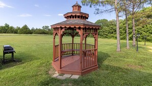 A gazebo with a wooden structure and a roof, surrounded by a grassy field and trees.