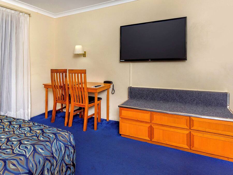 A hotel room with a bed, a desk with a chair, a TV mounted on the wall, and a wooden dresser.