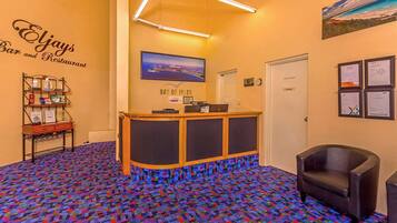 A reception area with a patterned carpet, a reception desk, a wall-mounted TV, and a framed picture of a landscape.