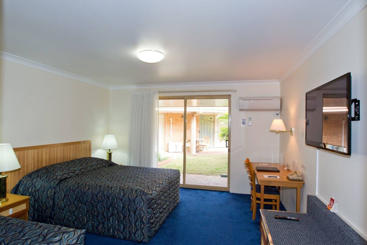 Une chambre d’hôtel équipée d’un lit, d’un bureau, d’une chaise, d’une télévision et d’une porte-fenêtre coulissante donnant sur un espace extérieur.