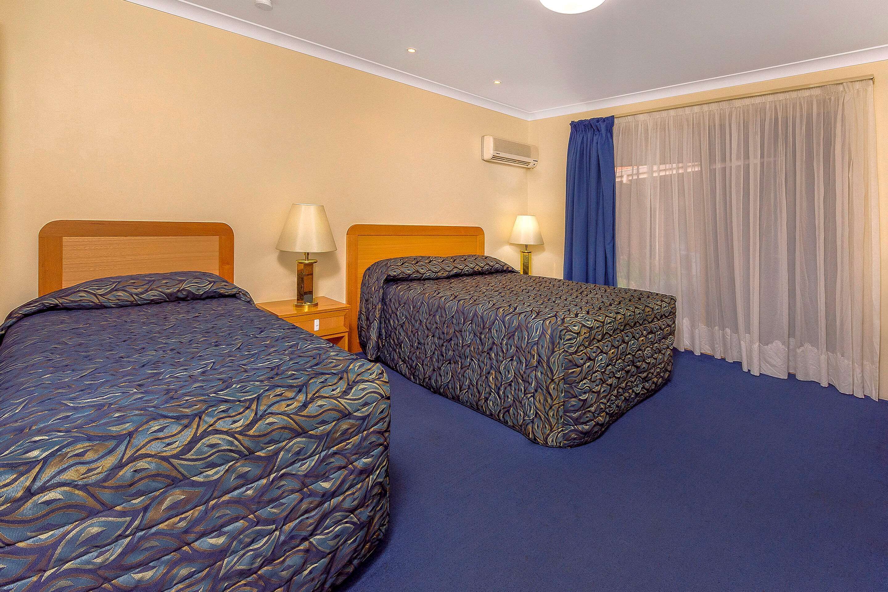 Une chambre d’hôtel avec deux lits, une moquette bleue et une fenêtre ornée de rideaux légers.