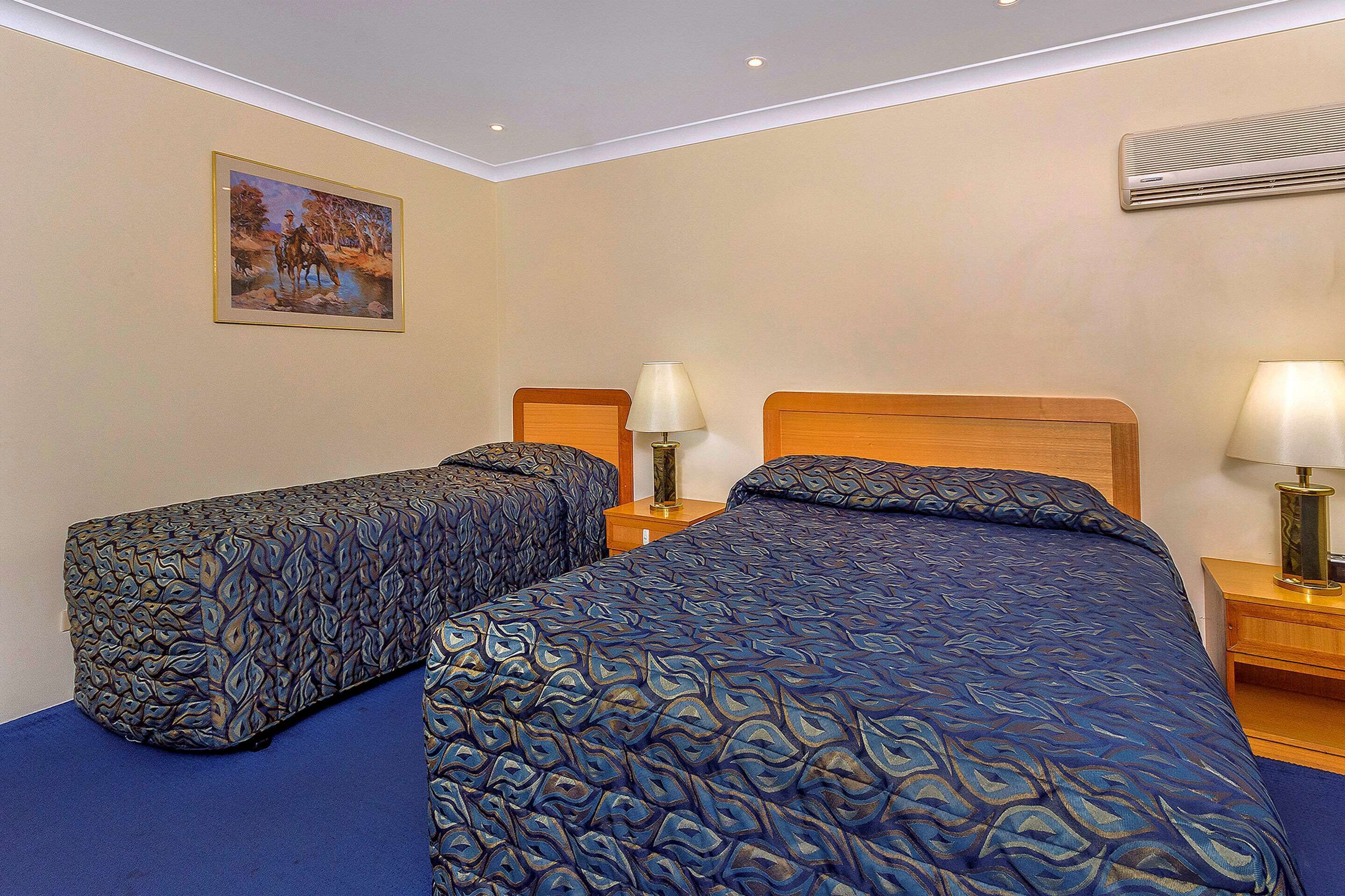 A hotel room with two beds, a nightstand with lamps, a painting on the wall, and an air conditioning unit.