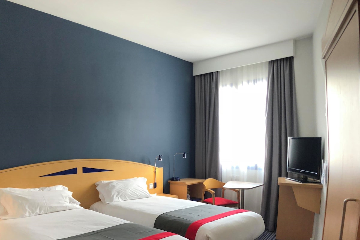 Hotellihuone, jossa on kaksi sänkyä, työpöytä, tuoli, televisio ja ikkuna, jossa on verhot.