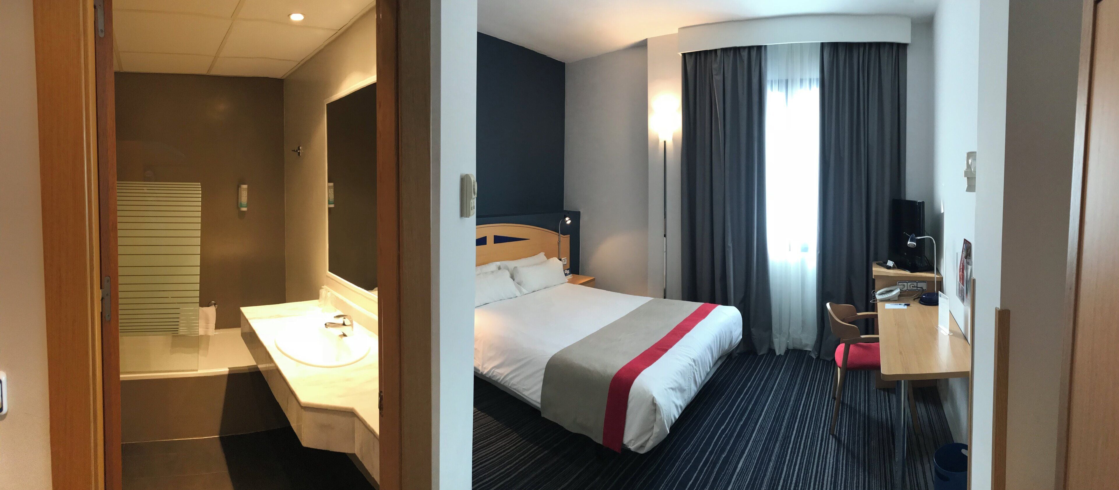 A hotel room with a bed, a desk, a chair, a desk lamp, and a television.
