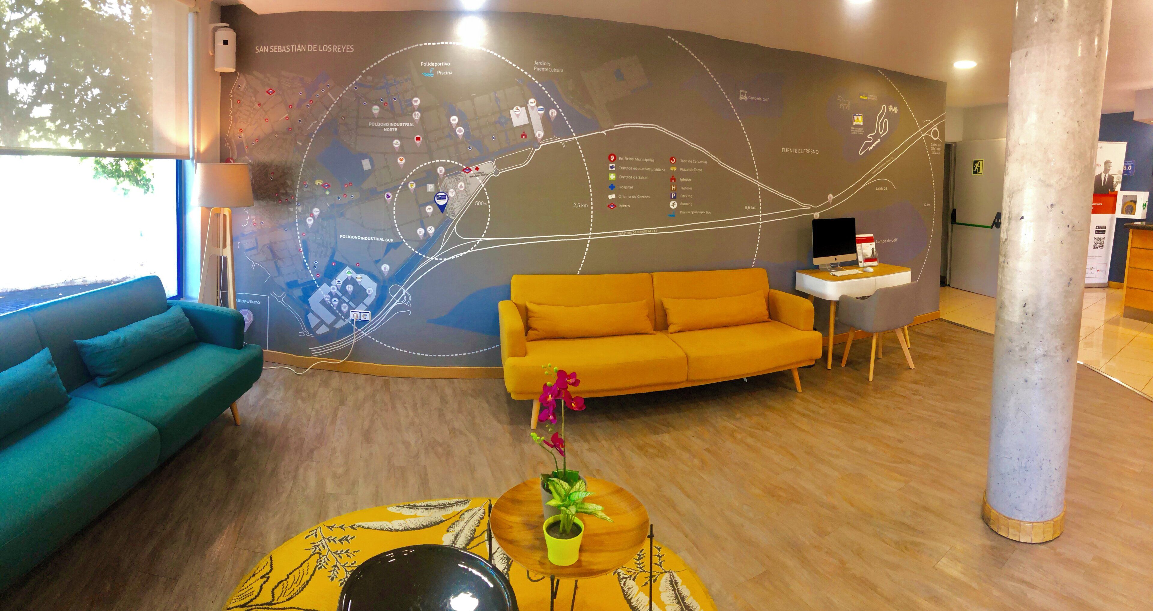 Un interior moderno con un gran mapa mural, un sofá verde azulado, un sofá amarillo, una mesita con una planta y un escritorio con una computadora.