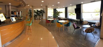 Un espacio interior amplio con una recepción, varios conjuntos de asientos y grandes ventanales que aportan luz natural.