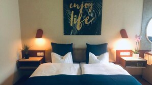 Uma cama bem arrumada com roupa de cama branca e azul, duas mesas de cabeceira com abajures e uma obra de arte na parede com a inscrição 'enjoy life'.
