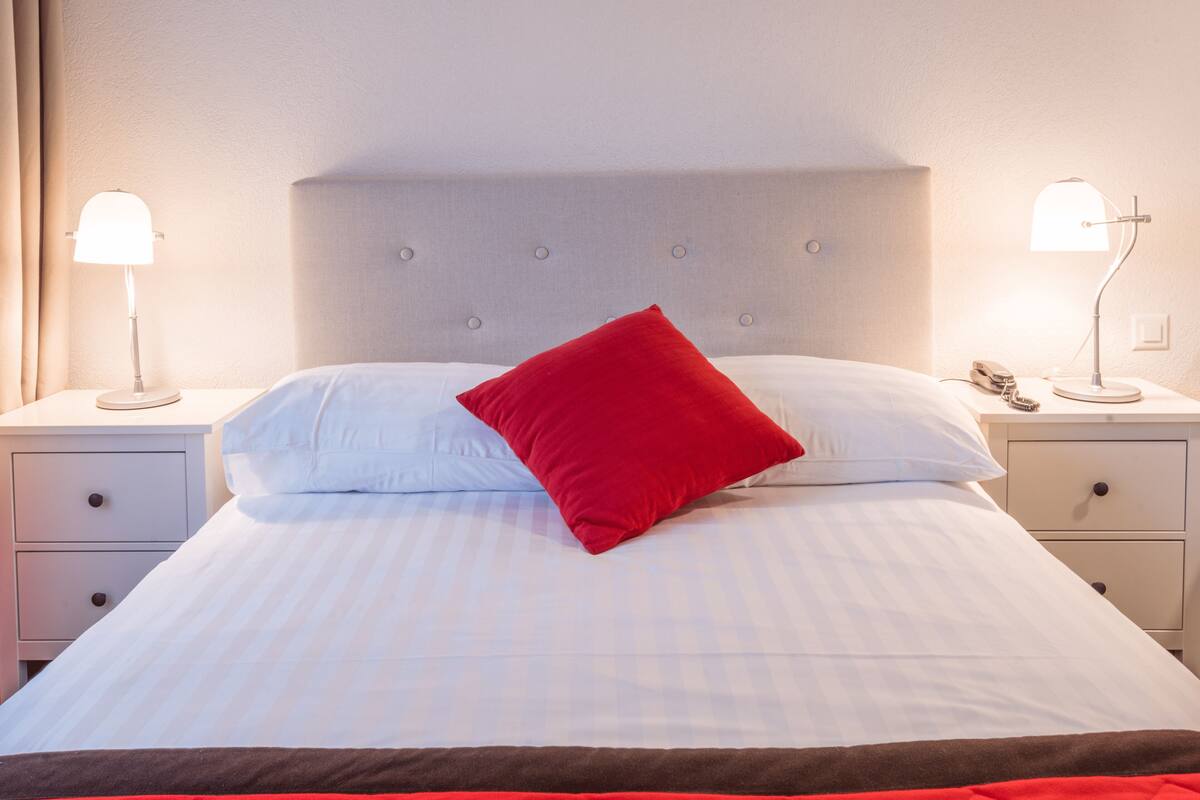 A neatly made bed with a red pillow, two bedside tables with lamps, and a headboard.