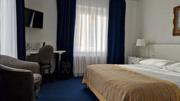 Habitación de hotel con cama, escritorio, silla, televisor y ventana con cortinas.