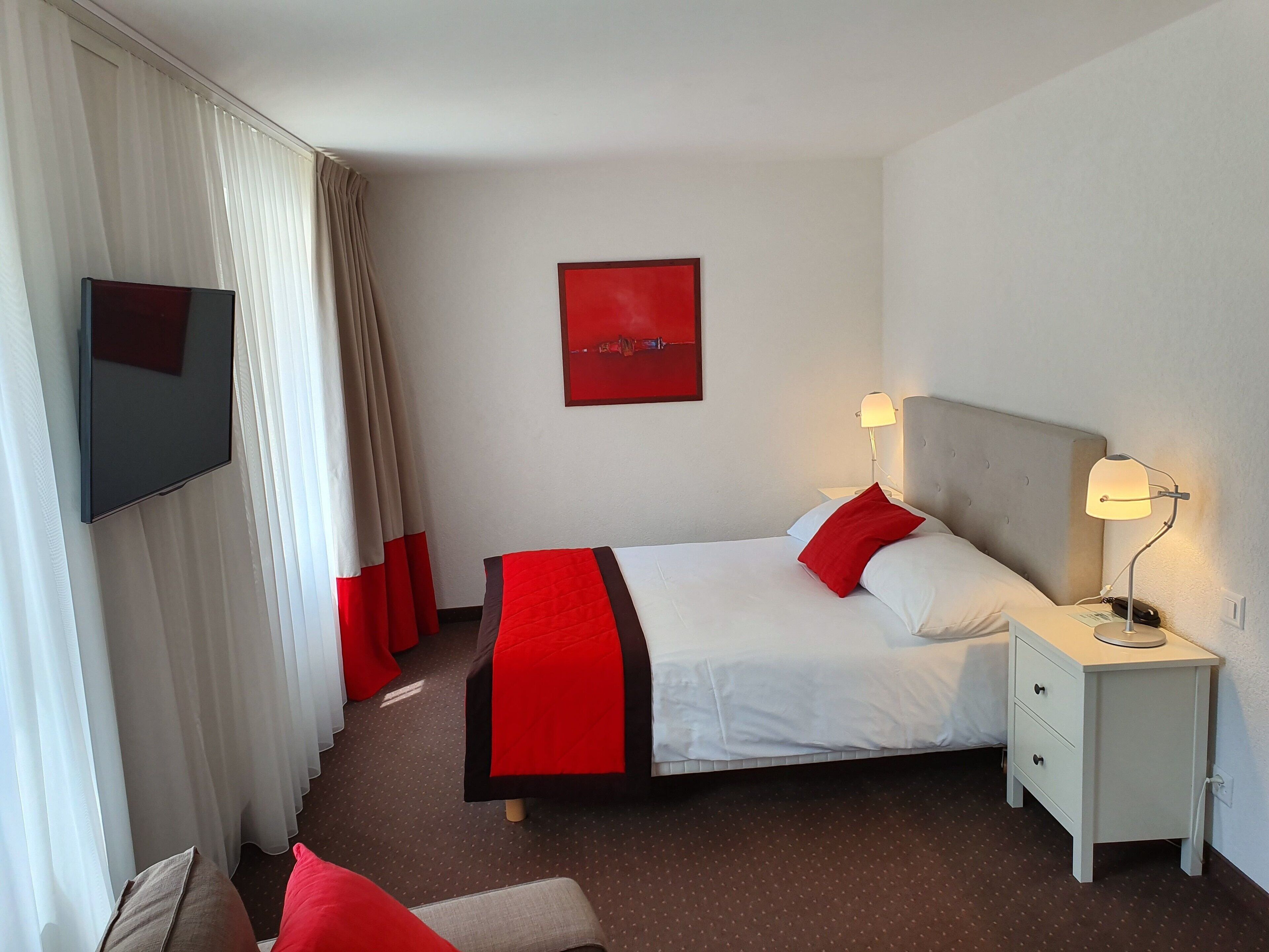 Ein Hotelzimmer mit einem Bett, einem an der Wand montierten Fernseher, einem Nachttisch mit Lampe und einem roten Kunstwerk an der Wand.