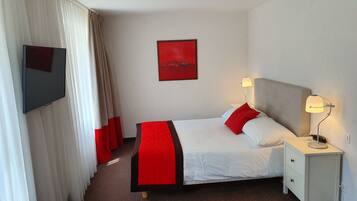 A hotel room with a bed, a TV mounted on the wall, a bedside table with a lamp, and a red artwork on the wall.