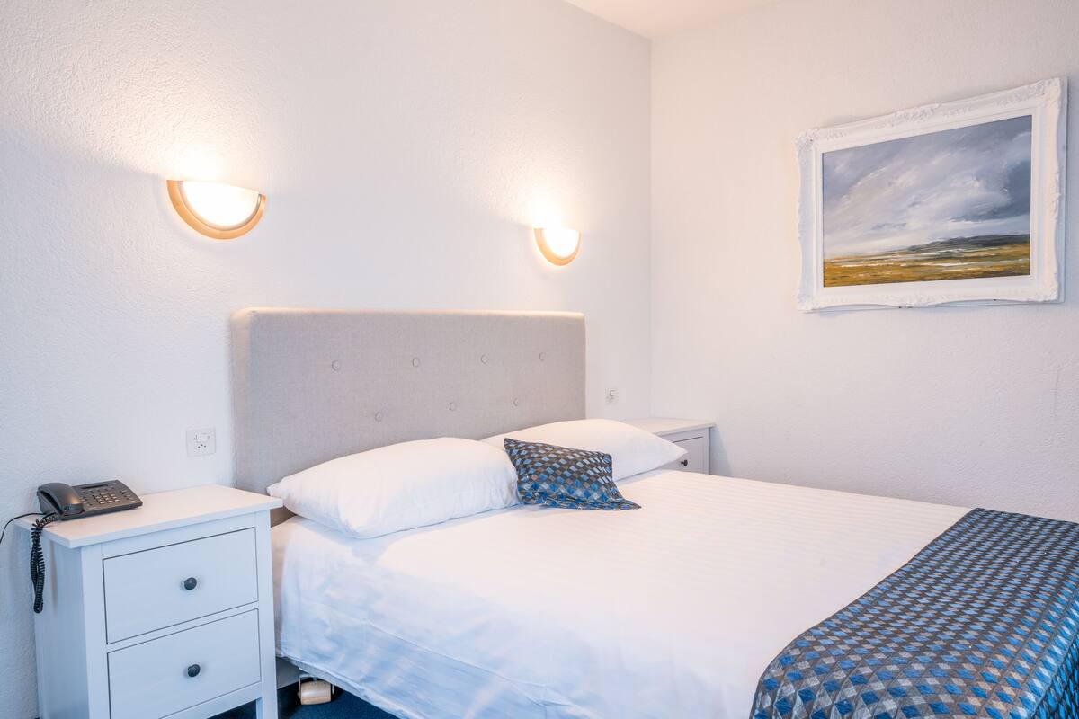 A neatly made bed with white linens and a patterned throw, a bedside table with a phone, and two wall-mounted lights.