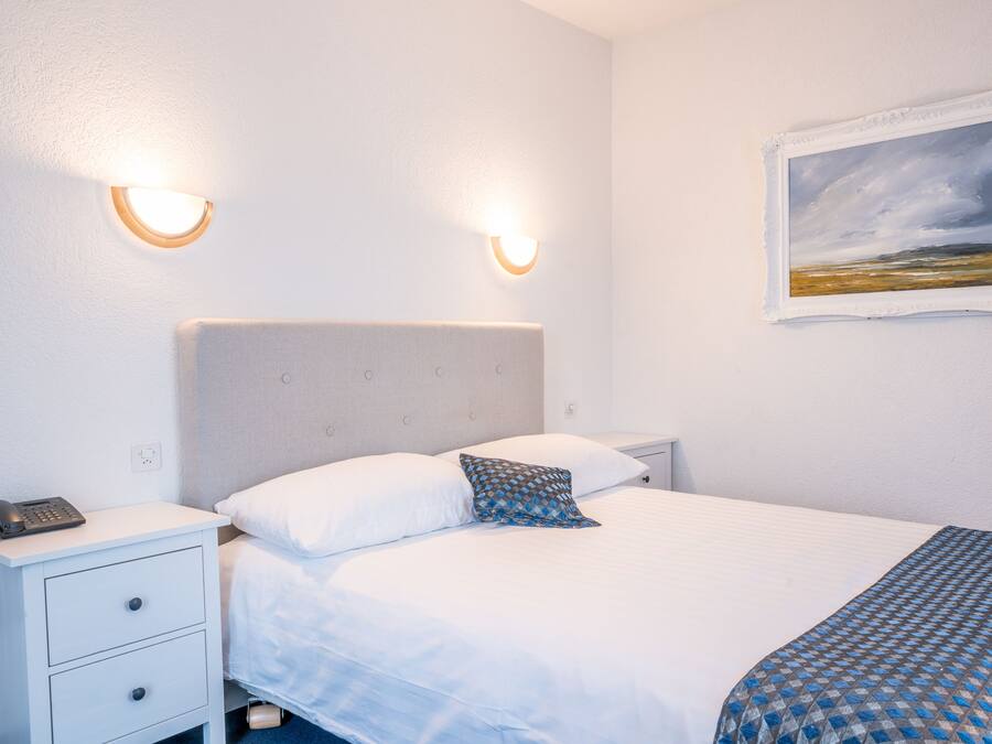 Un lit bien fait, avec du linge de lit blanc et un couvre-lit à motifs, une table de chevet avec un téléphone et deux lampes fixées au mur.
