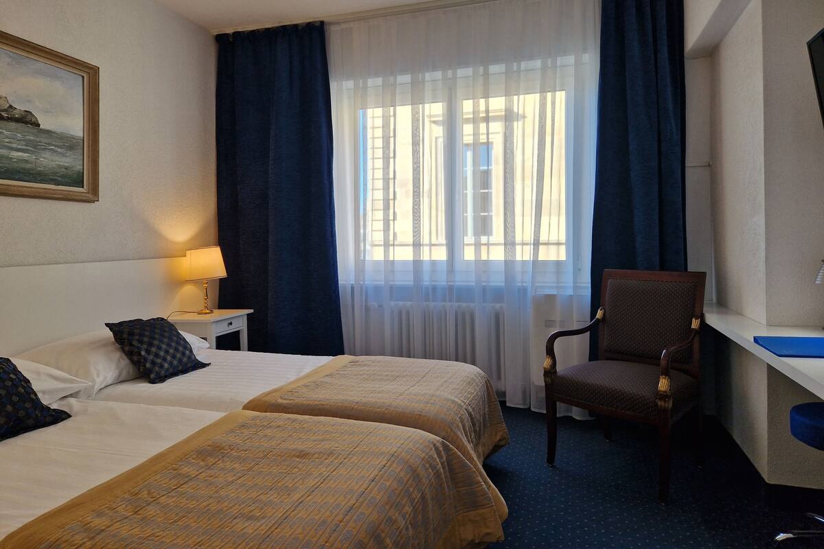 A hotel room with two beds, a desk, a chair, a lamp, and a window with curtains.