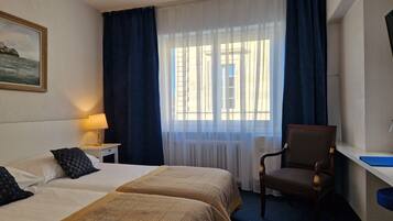 Habitación de hotel con dos camas, un escritorio, una silla, una lámpara y una ventana con cortinas.