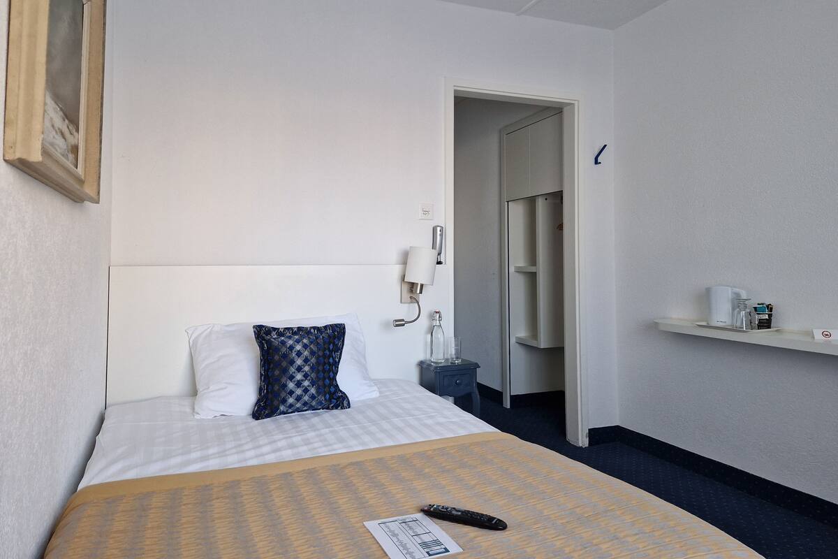 A hotel room with a bed, a bedside table, a nightstand, a wall-mounted lamp, a shelf with toiletries, and a door leading to another room.