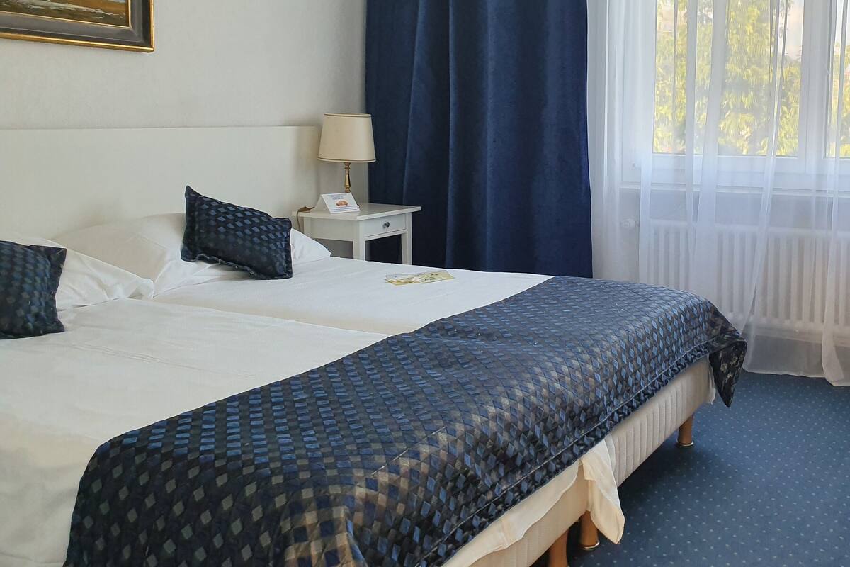 A hotel room with two beds, a nightstand, a lamp, a framed picture on the wall, and a window with curtains.
