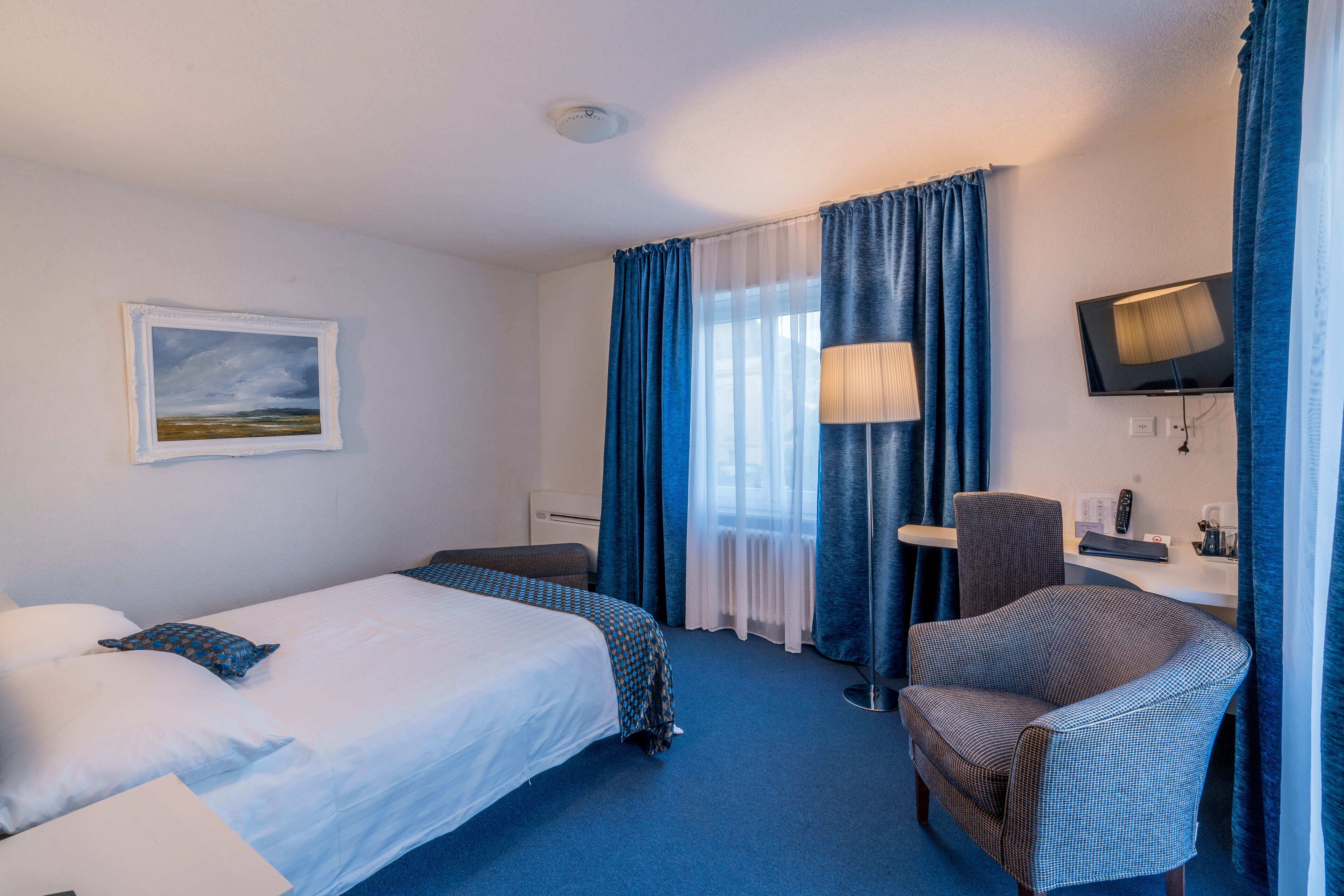 A hotel room with a bed, a chair, a desk with a lamp, a television, and a painting on the wall.